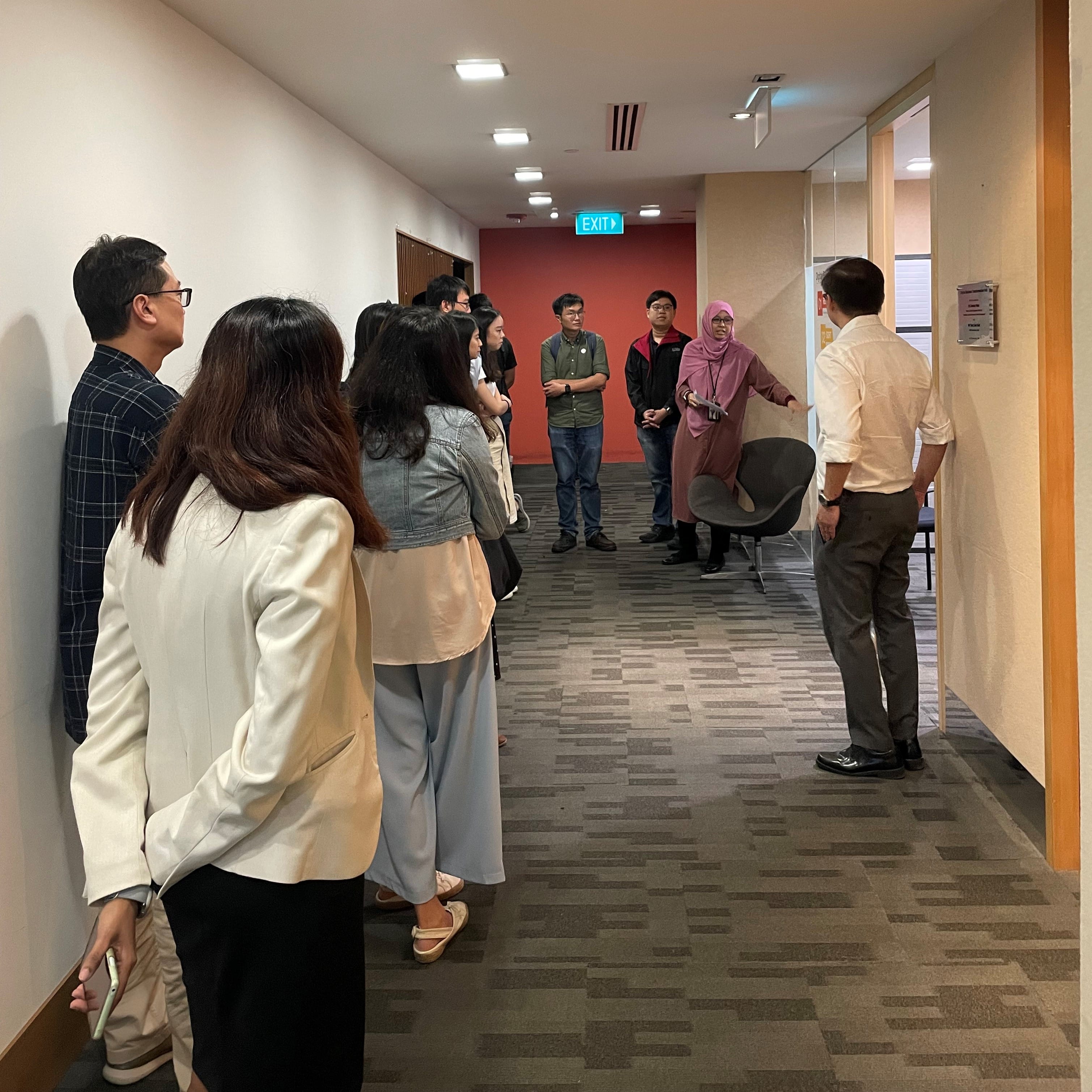 A group of about ten people standing in a carpeted hallway near an exit sign, listening to a man in a white shirt.